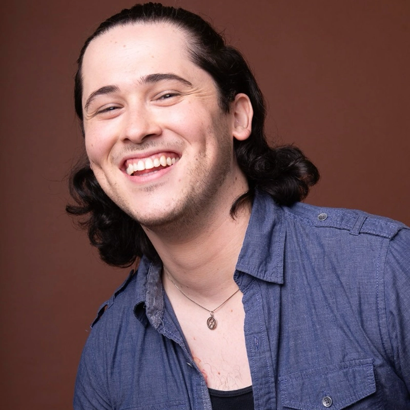 Zachary Santolaya, a smiling person with long, dark, wavy hair pulled back, wears a blue button-up shirt over a black top and a necklace while posing in front of a plain brown background.