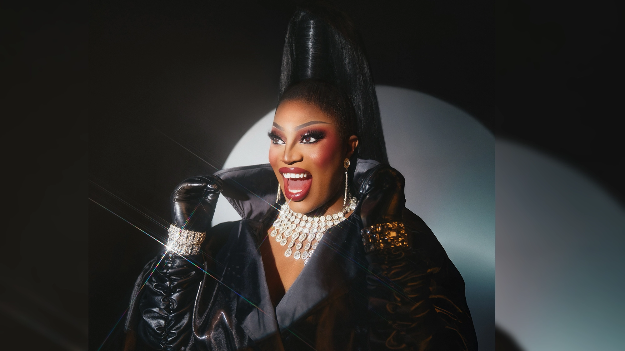 A drag queen, reminiscent of Sapphira Cristál in a shiny black outfit, large gloves, sparkling jewelry, and dramatic makeup, smiles with their collar raised. Their hair is styled in a tall, sleek updo against a dark background with a white circular light behind them—radiating the fierce energy of “Sapphi Don’t Sing The Blues.”.