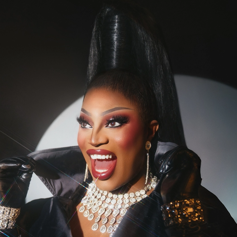 A person with dramatic makeup, long black hair styled high, wearing black gloves, a dark outfit, sparkling jewelry, and earrings, smiles widely against a dark background with a circular light spot.