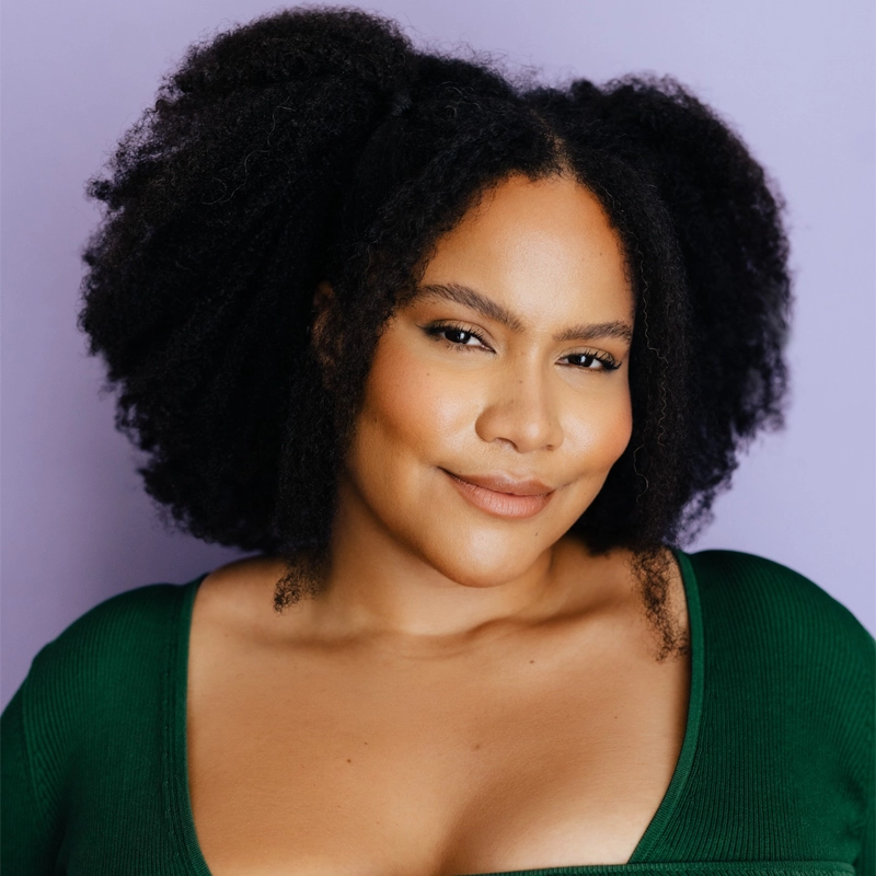 Ashley Seldon, a woman with voluminous natural curly hair, smiles softly at the camera. She wears a deep green top with a low neckline. Her makeup is natural, with bronzed cheeks and nude lipstick. The background is a solid pale lavender color.