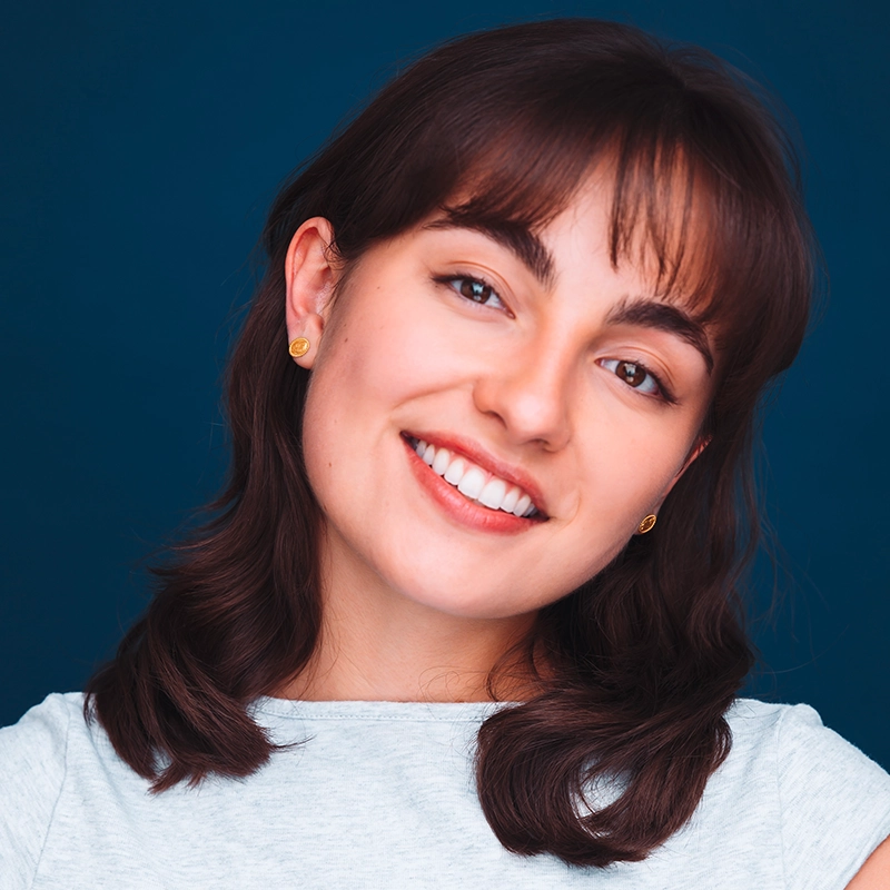 A young woman, Phoebe Kleehammer, with shoulder-length dark brown hair smiles at the camera, wearing a light gray top and small gold earrings, against a dark blue background. Her head is slightly tilted and her teeth are showing.