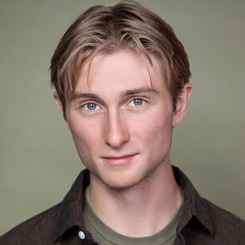 A young adult with fair skin, light brown hair parted in the center, and blue eyes looks directly at the camera with a slight smile. Spencer Bowman wears a dark shirt over a green T-shirt. The background is plain and muted green.