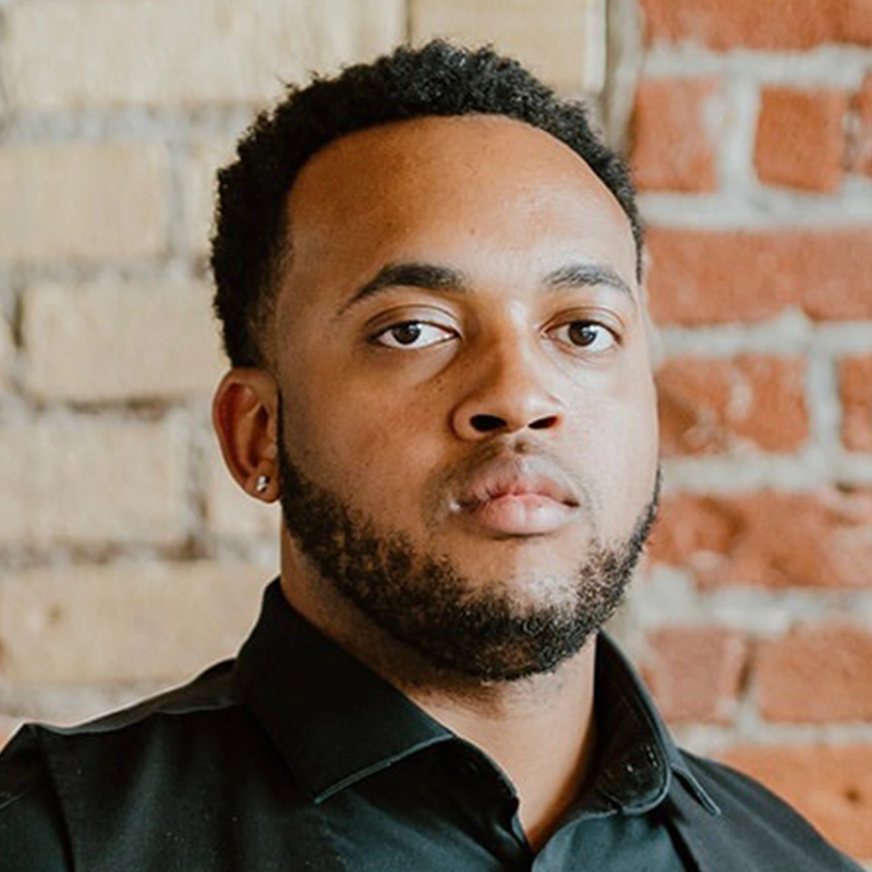 A man with short curly hair, trimmed beard, and mustache wears a black collared shirt. He has a neutral expression, looking directly at the camera. The background shows exposed red and beige brick walls, creating a distinctive setting for Andre Cleghorn’s portrait.
