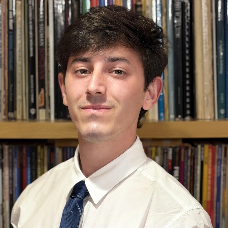 A young man with short dark hair, wearing a white dress shirt and blue tie, stands in front of shelves filled with colorful books or records. Kosta Stoychev looks at the camera with a slight smile and a neutral expression.