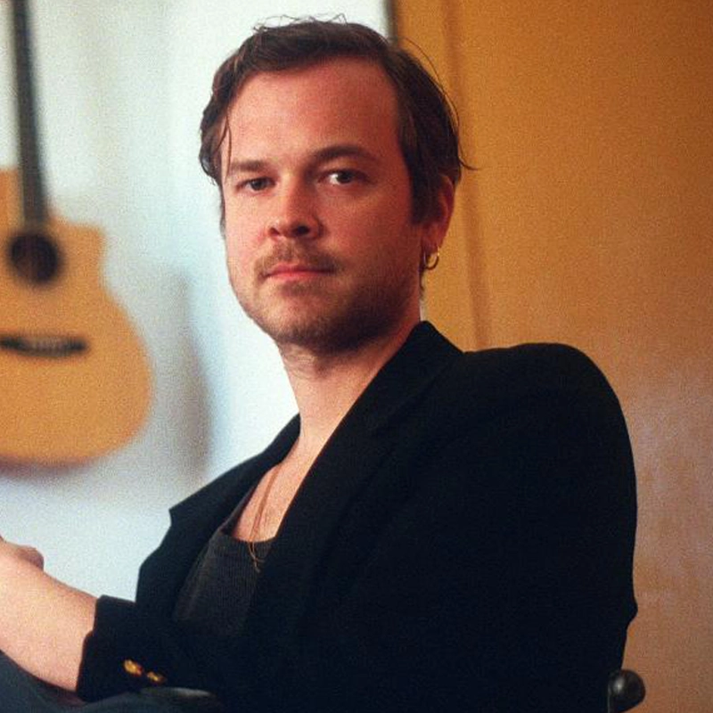 A man with short brown hair and light facial hair sits indoors, wearing a black jacket and a thin necklace. He looks calmly at the camera. A wooden acoustic guitar hangs on a yellow wall behind him, and the lighting is soft and warm.