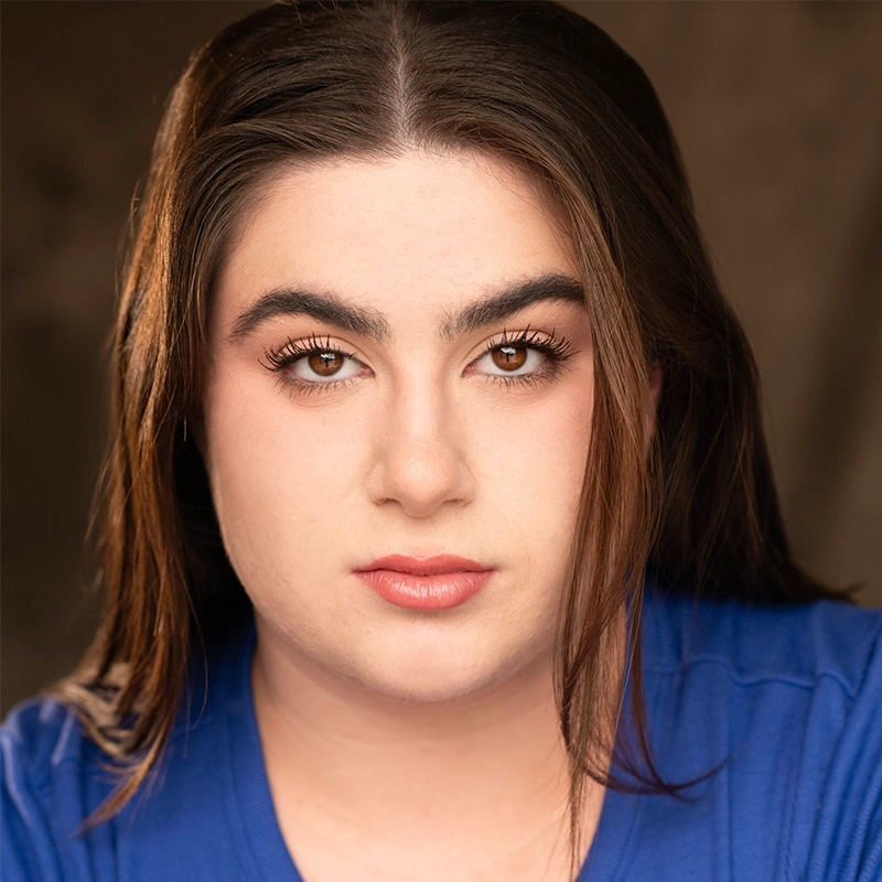 Spencer Bowman, a person with long brown hair and thick eyebrows, looks directly at the camera with a neutral expression. They have fair skin, brown eyes, and wear a blue shirt. The background is softly blurred in warm brown tones.