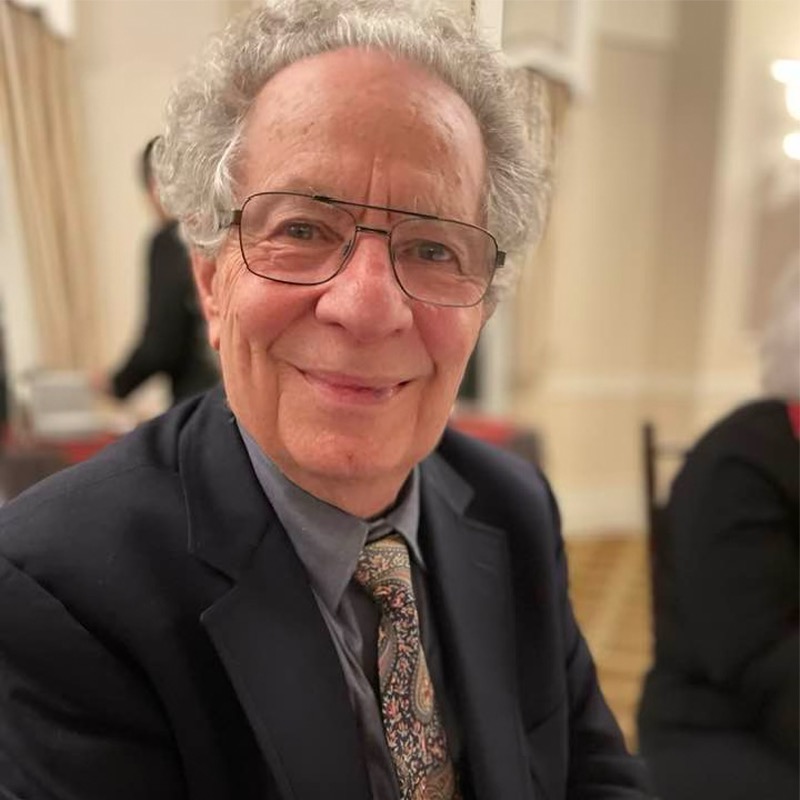 An older man with curly gray hair and glasses, wearing a dark suit, patterned tie, and light shirt, smiles warmly at the camera while seated indoors at a formal event. The background is softly blurred, showing curtains and another person. This distinguished gentleman is Thomas Z. Shepard.
