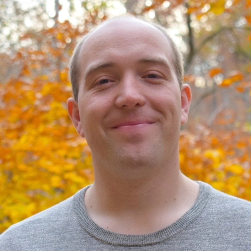 A man with light skin and short brown hair smiles gently outdoors. He wears a light gray crewneck sweater. The background is blurred, showing trees with autumn leaves in vibrant yellow, orange, and hints of green. The lighting is soft and natural, evoking the serene mood often captured in Melina Walko’s photography.
