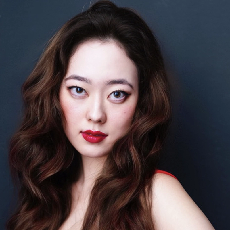 Katherine Vandermel, a woman with long, wavy brown hair and fair skin, poses against a dark background. She wears bright red lipstick, subtle makeup, and a sleeveless red top, looking directly at the camera with a confident, neutral expression.