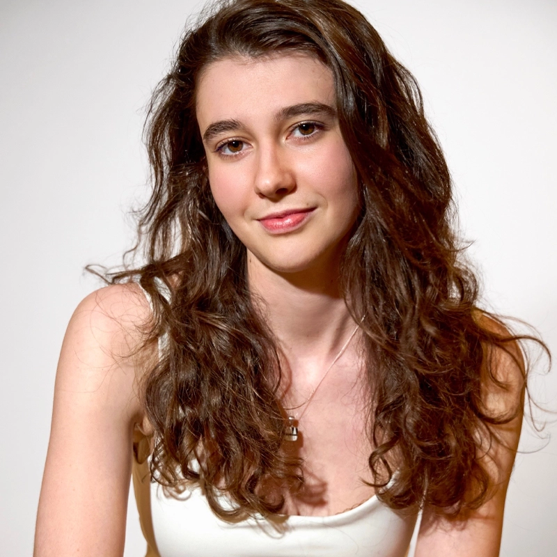 A young woman named Samantha White with long, wavy brown hair and fair skin sits against a plain white background, wearing a white tank top and a necklace. She smiles softly, looking directly at the camera with a relaxed and confident expression.
