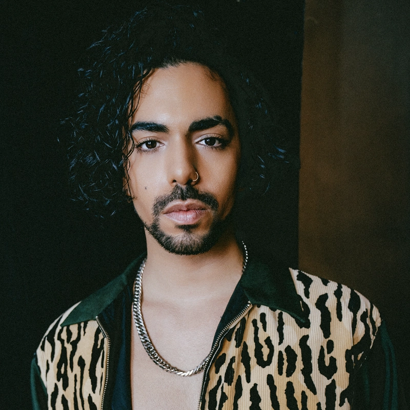 A portrait of Maximo Xtravaganza with dark, curly hair, a beard, and a nose ring, looking directly at the camera. They are wearing a black shirt under a tan and black patterned jacket with a wide collar and a thick silver chain necklace, set against a dark background.