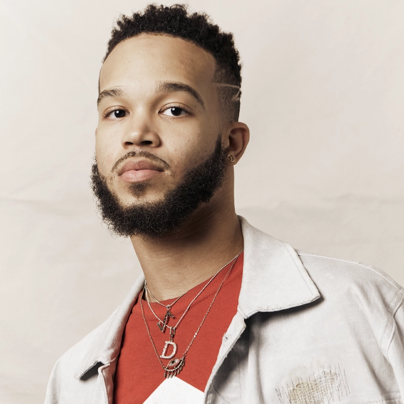 A young man with a faded haircut and groomed beard looks toward the camera, sporting a legendary look in a light denim jacket and red shirt. Layered silver necklaces complete the style against a neutral cream background.
