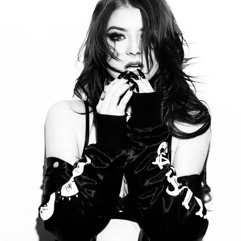 A young woman with long, dark hair poses against a white wall, looking directly at the camera with her fingers near her lips. She wears a black, long-sleeved jacket with decorative white patterns on the sleeves and has dark, manicured nails. The image is in high-contrast black and white.
