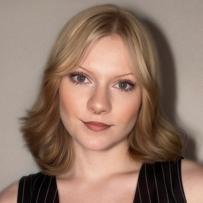 A young woman with light skin and shoulder-length blonde hair styled in loose waves looks at the camera. She has green eyes, subtle makeup, and wears a black sleeveless top with thin white stripes. Corinne Mickey Durkin has a septum piercing and a neutral expression against a plain background.