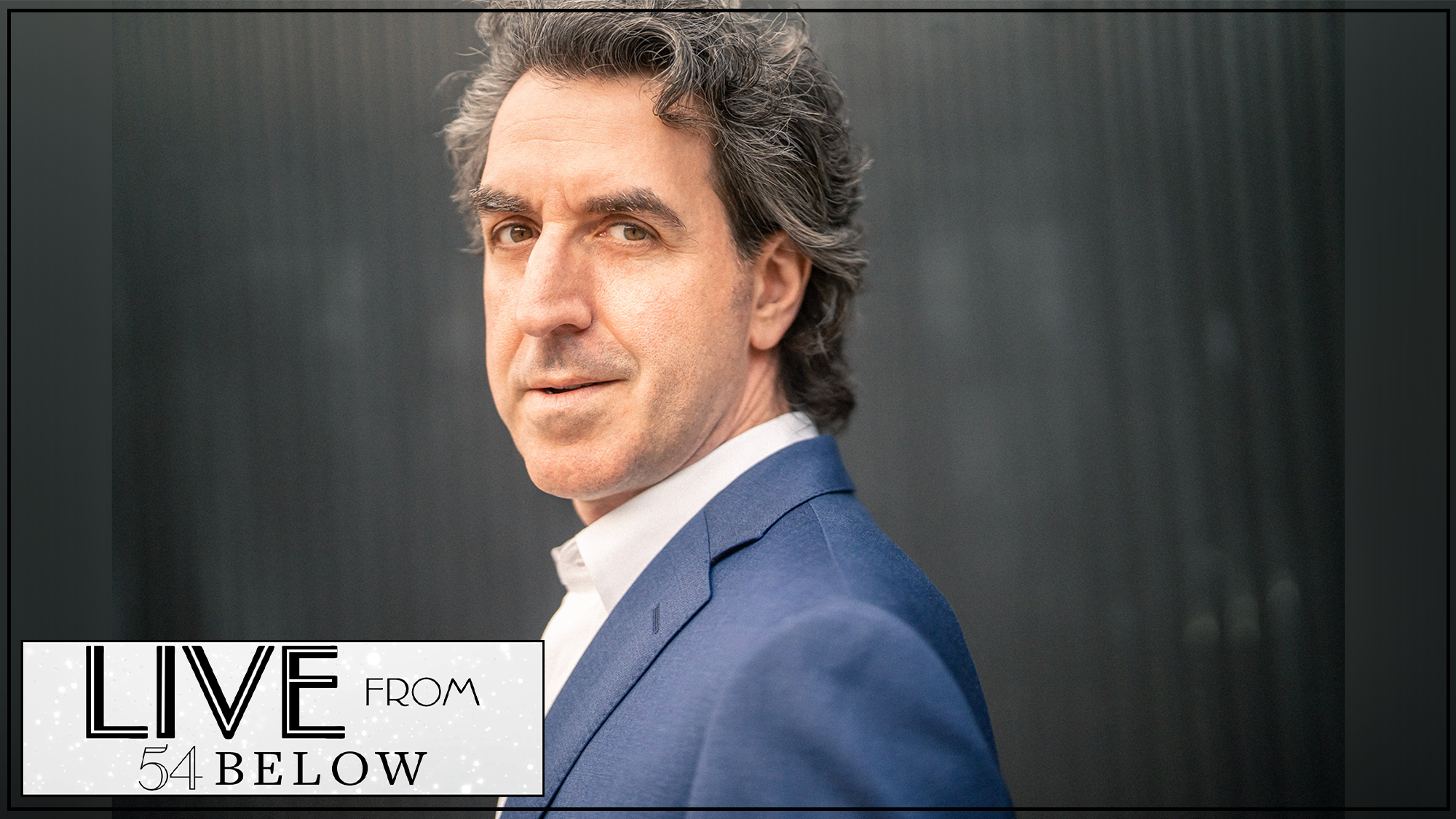 A man with wavy graying hair wears a blue suit and white shirt, looking over his shoulder at the camera. The background is dark and blurred. In the bottom left corner, a box reads “LIVE FROM 54 BELOW” in bold text, highlighting an NYC LIVESTREAM event.