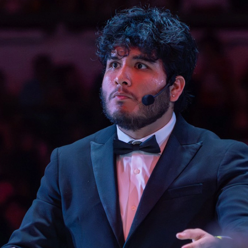Juan Aguirre, a man with dark curly hair and a beard, looks intensely to the side while wearing a black tuxedo, white shirt, and bow tie. He wears a small microphone headset and is captured from the chest up against a dimly lit background.