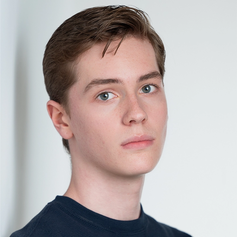 A headshot of Ryan Bagnolati-Camargo, a young person with short brown hair and light eyes looking directly at the camera. They have a neutral expression and are wearing a dark blue crew-neck shirt against a plain white background.