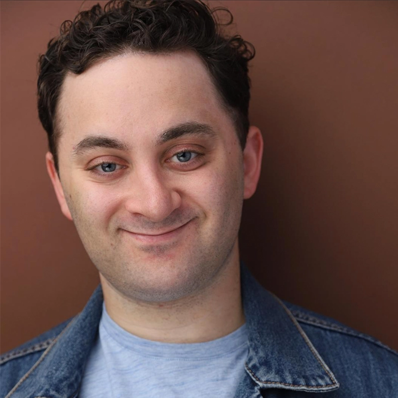 A headshot of a man with curly brown hair and blue eyes, smiling slightly at the camera. He is wearing a light blue t-shirt under a denim jacket against a solid brown background.