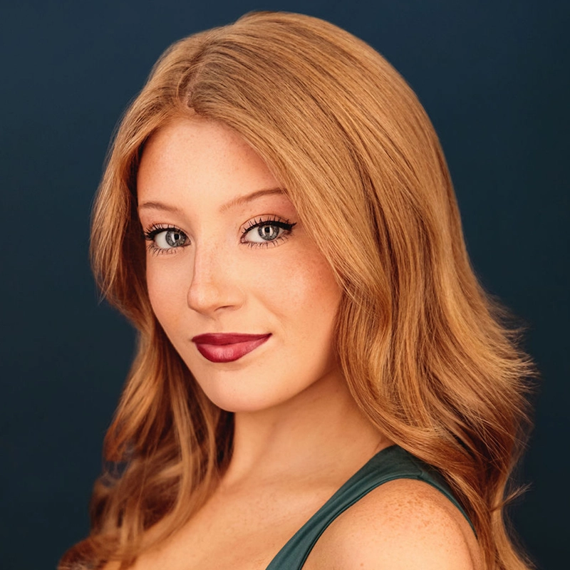 Ryan Bagnolati-Camargo is a young woman with long, wavy auburn hair and freckles, smiling at the camera against a dark blue background. She wears dark lipstick and a deep green top, with her hair parted in the center and framing her face.
