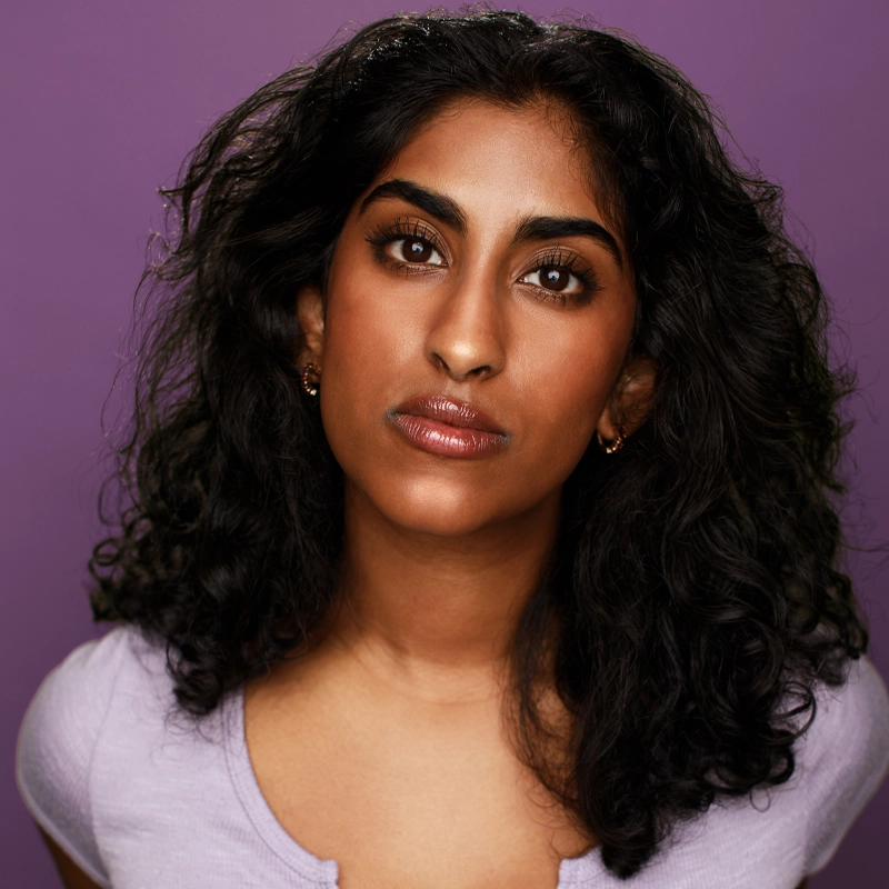 A portrait of Hashini Amarasinghe with long, dark, wavy hair and a neutral expression against a purple background. She is wearing a light purple scoop-neck shirt and small gold hoop earrings, looking directly at the camera.