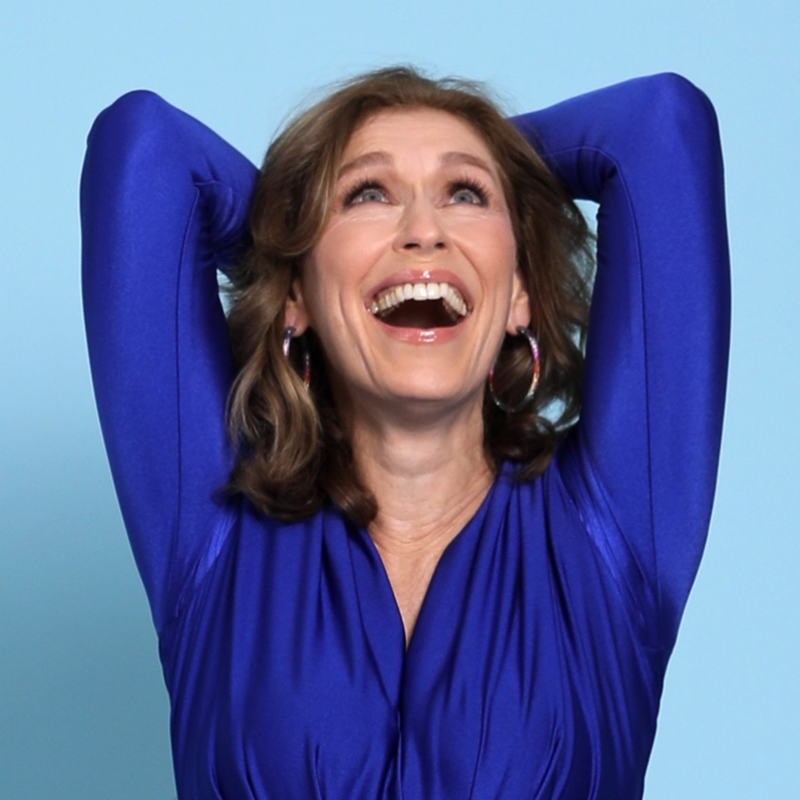 A woman with shoulder-length brown hair laughs with her head tilted back and her arms raised behind her head. She is wearing a bright blue long-sleeved dress and colorful hoop earrings against a light blue background.