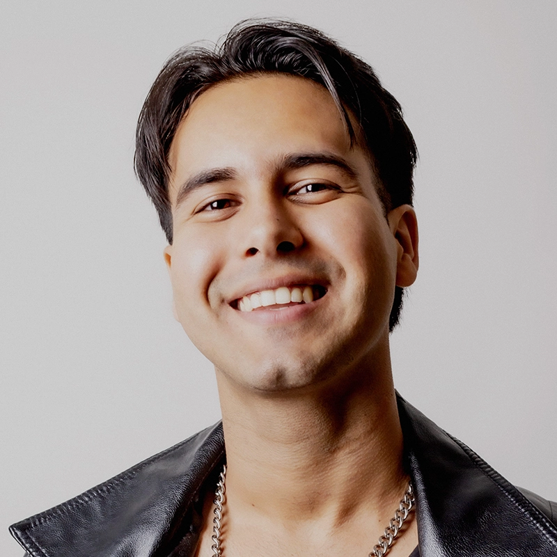 Ryan Bagnolati-Camargo smiles broadly at the camera. He has dark, styled hair and wears a black leather jacket over a dark top with a silver chain necklace, set against a plain, light gray background.