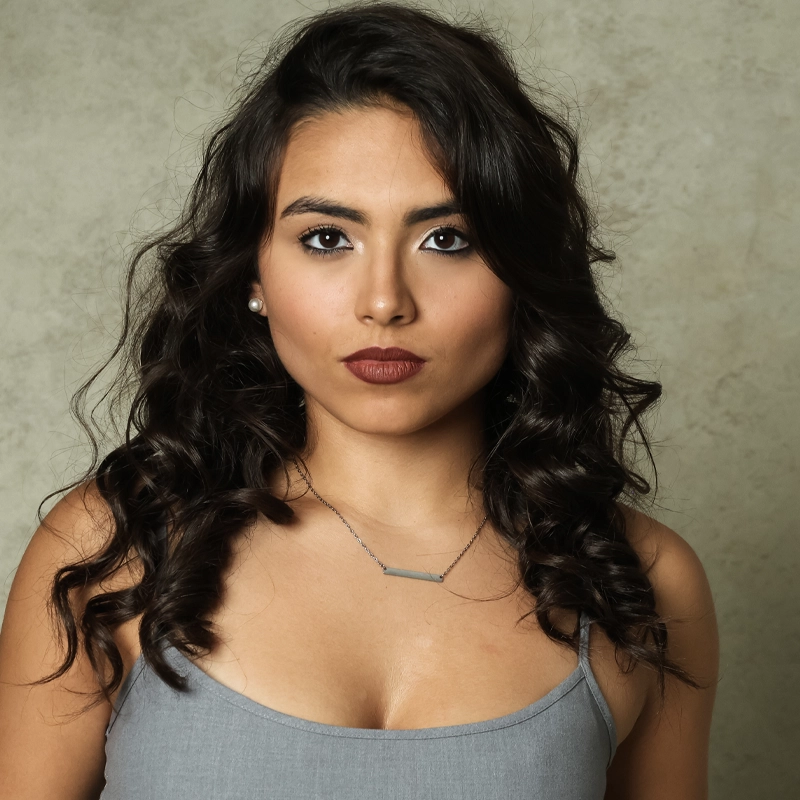 A head-and-shoulders portrait of Arianna Arocho, a young woman with long, dark wavy hair, brown eyes, and dark lipstick. She is wearing a grey tank top, a small silver bar necklace, and simple pearl stud earrings against a textured, light-grey background.
