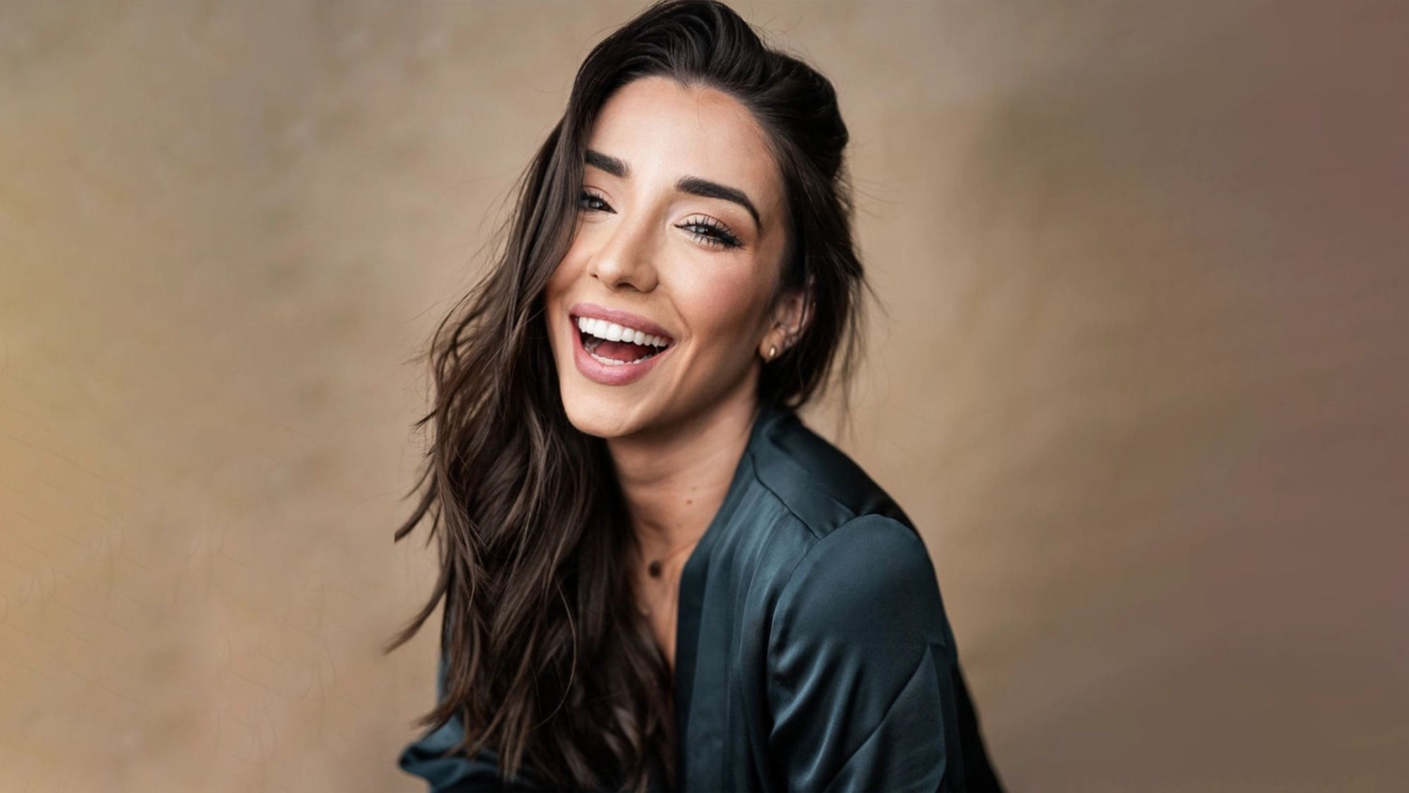 Ashley Loren smiles at the camera, her long dark hair flowing over a dark green top against a neutral background.