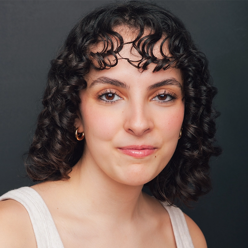 A young person with short, dark, curly hair and finger waves on their forehead smiles softly. Bella Brodine wears a sleeveless light top and gold hoop earrings, posed against a plain, dark background with a calm, confident expression.