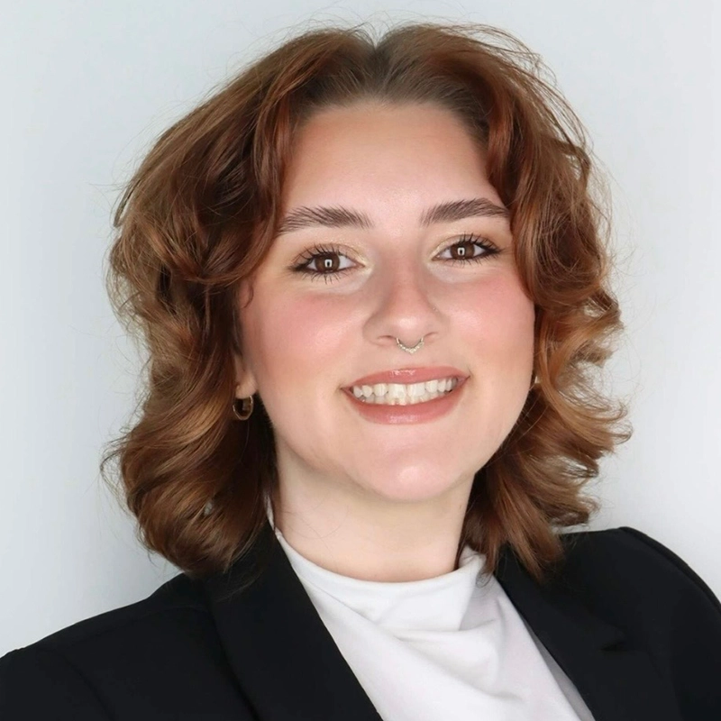 Smiling person with fair skin, wavy shoulder-length brown hair, and a septum piercing. They wear hoop earrings, a white high-neck top, and a black blazer, posing against a plain light background.
