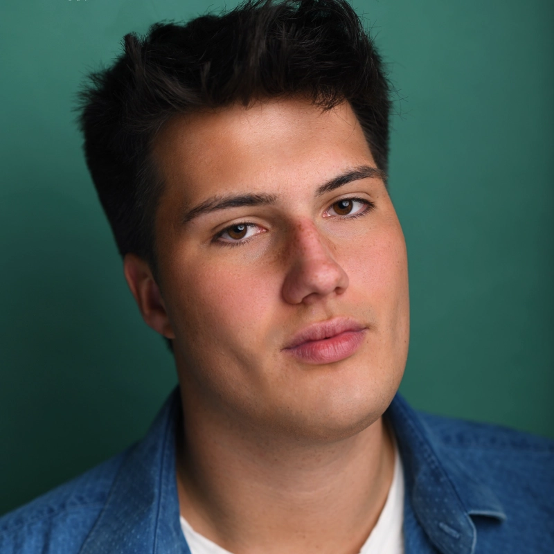 Brandon Rundquist, a young man with dark hair, looks toward the camera against a green background. He is wearing a blue denim shirt over a white undershirt.