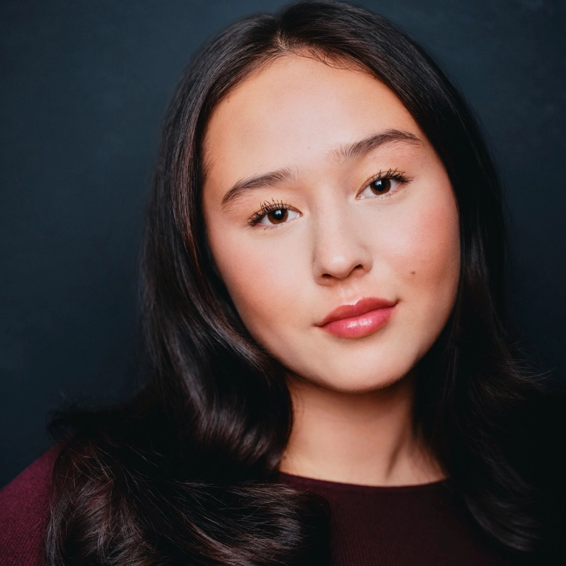 Carolyn Burch is a young woman with long, dark wavy hair and a neutral expression, facing the camera against a dark background. She wears a dark burgundy top, with her hair parted in the center to frame her face.