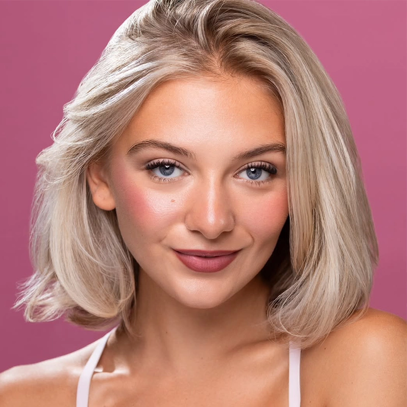 A young woman with shoulder-length, platinum-blonde hair smiles softly at the camera. She has light skin, blue eyes, and rosy cheeks, posed against a solid, muted pink background.