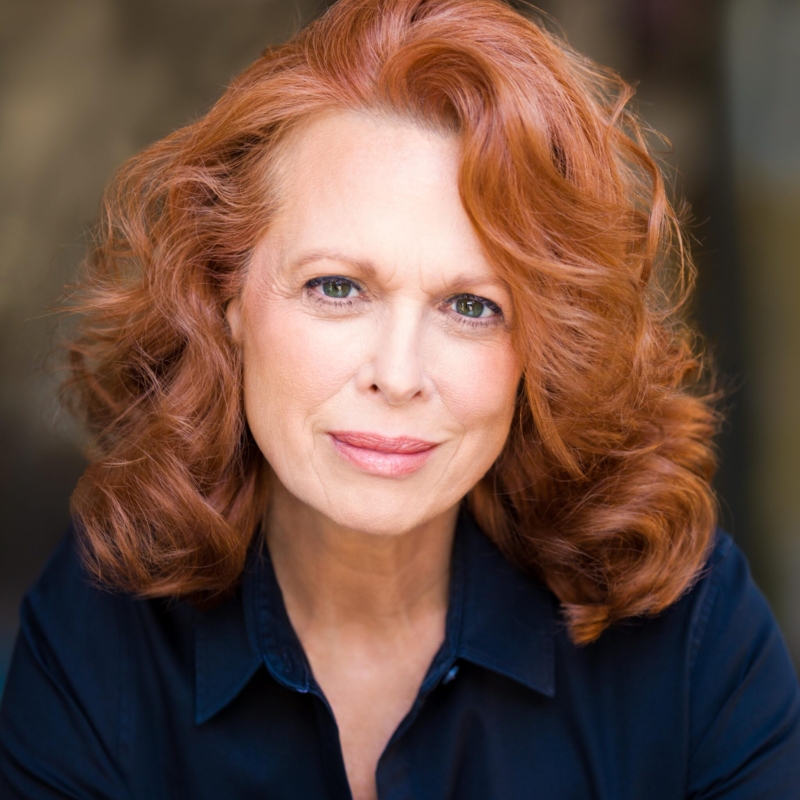 A headshot of a middle-aged woman with wavy auburn hair looking directly at the camera. She is wearing a dark collared shirt.