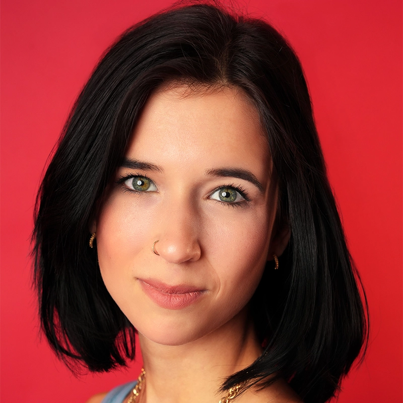 A portrait by Tyler Cirulnick of a young woman with dark hair, green eyes, and a nose ring, shown from the head and shoulders against a solid red background.