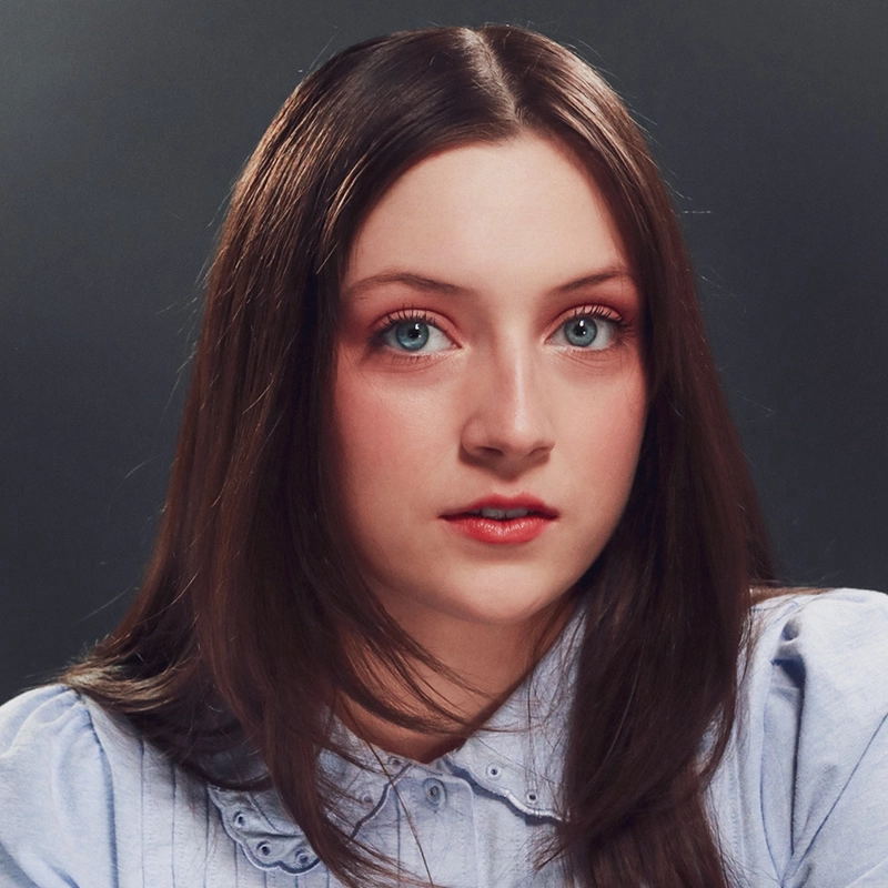A head-and-shoulders portrait of Tyler LaPointe, a young woman with long, dark brown hair and bright blue eyes looking directly at the camera. She wears a light blue collared shirt and has soft pink eyeshadow and lip color, set against a dark, moody gray background.