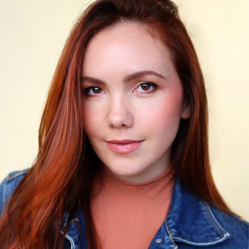 A young woman with long red hair looks toward the camera. She wears a coral-colored high-neck top under a denim jacket against a plain light background.