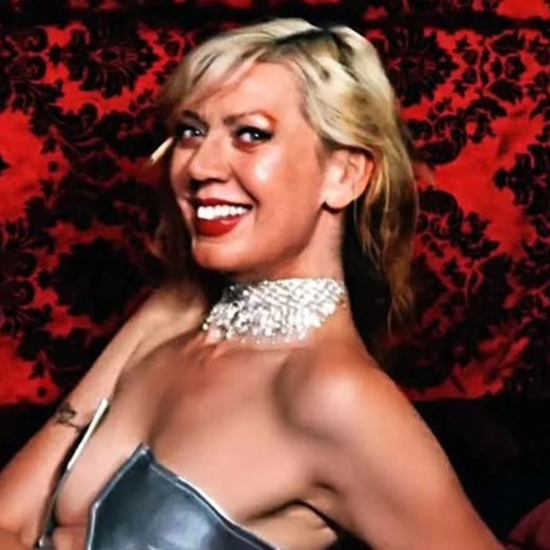 Charlene Hansen flashes a bright smile while wearing a shimmering, multi-layered rhinestone choker and a metallic silver top. She is positioned in front of a bold, deep red and black damask-patterned backdrop.