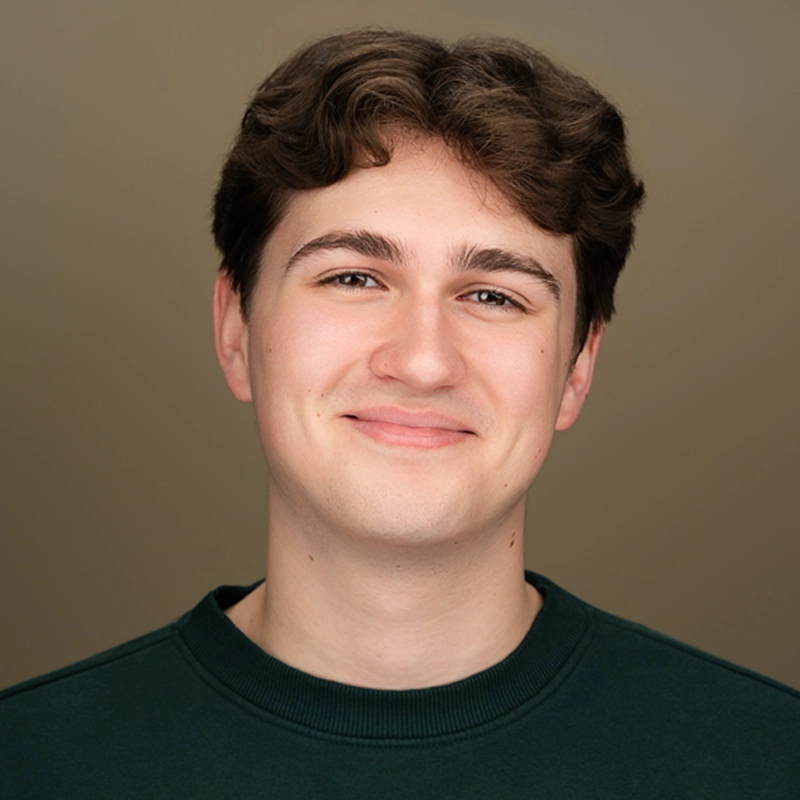 A portrait of Ryan Bagnolati-Camargo, a young man with wavy brown hair, looking directly at the camera with a gentle smile. He has light skin and is wearing a dark green crew-neck sweatshirt against a solid, muted brown background.