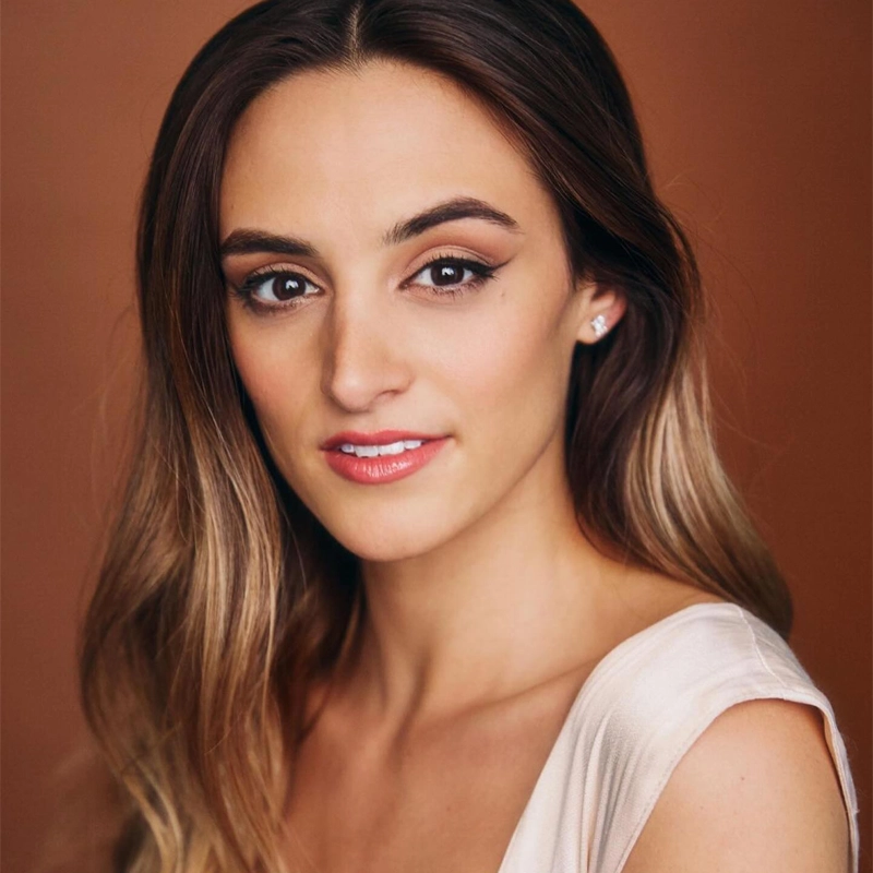 A headshot of a woman with long, wavy brown hair facing forward against a warm, solid rust-colored background. She has a neutral expression, winged eyeliner, and wears a light-colored sleeveless top and small stud earrings.