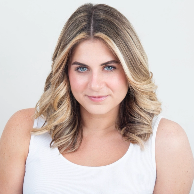 Leah Corcoran is a young woman with shoulder-length, blonde-highlighted hair and blue eyes looking toward the camera. She is wearing a simple white tank top against a plain, light-grey background.