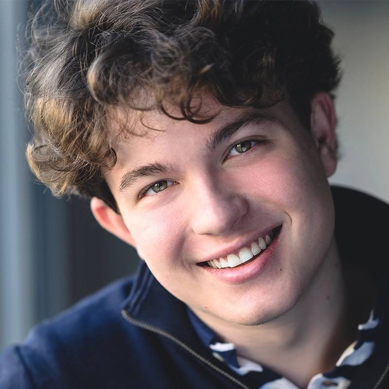 A close-up portrait of Ryan Bagnolati-Camargo, a young person with curly brown hair and green eyes, smiling warmly at the camera. They are wearing a dark blue zip-up sweater over a patterned collared shirt in soft, natural light against a neutral, blurred background.