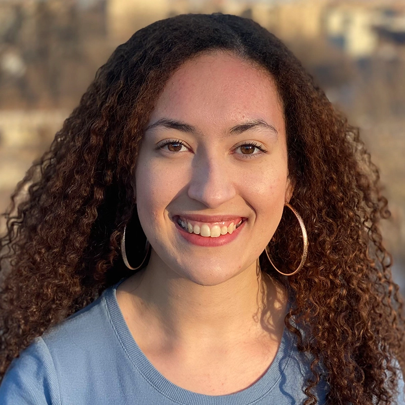 Tyler Cirulnick, a young woman with long curly hair and gold hoop earrings, smiles at the camera while wearing a light blue shirt.