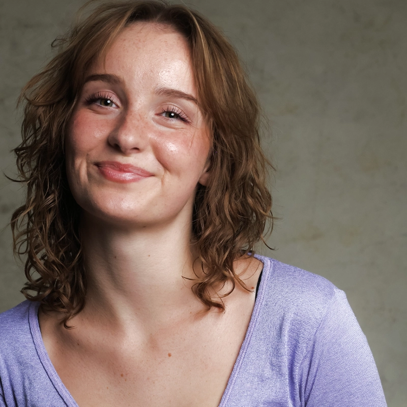 Abby DeGregory smiles warmly at the camera, her shoulder-length, wavy reddish-brown hair framing a face dotted with freckles. She wears a lavender scoop-neck top against a neutral, mottled gray background.
