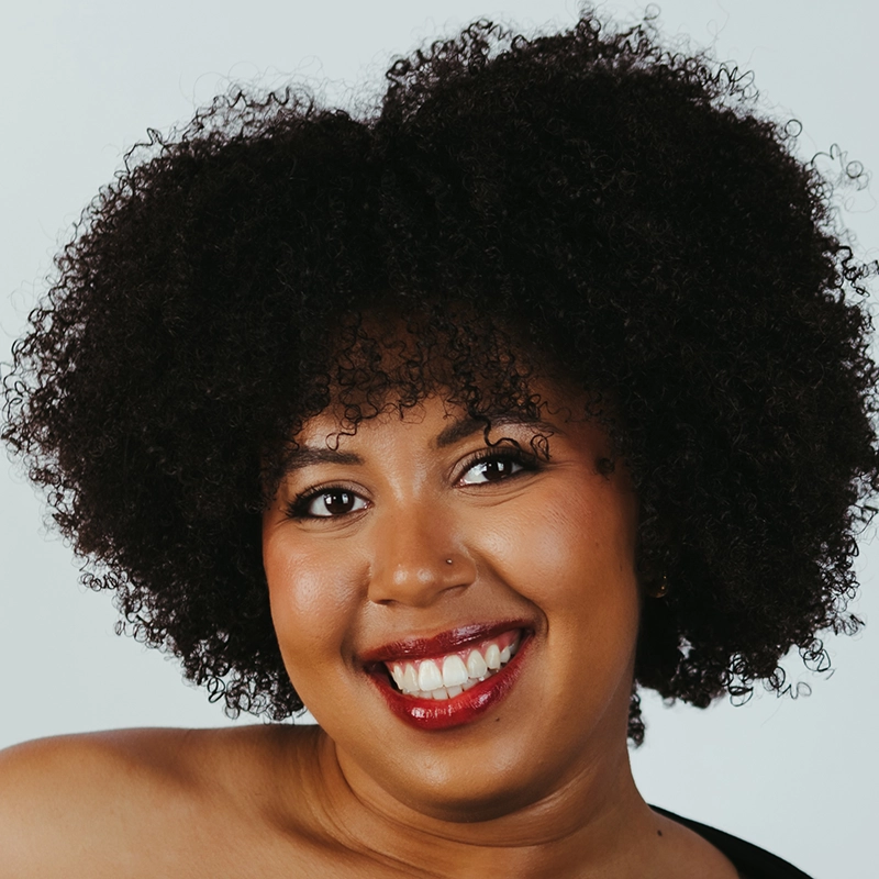A smiling woman with dark curly hair, brown eyes, and shiny red lipstick. She has smooth, glowing skin, a nose piercing, and wears a black top. The background is plain and light-colored. Her expression is warm and friendly.