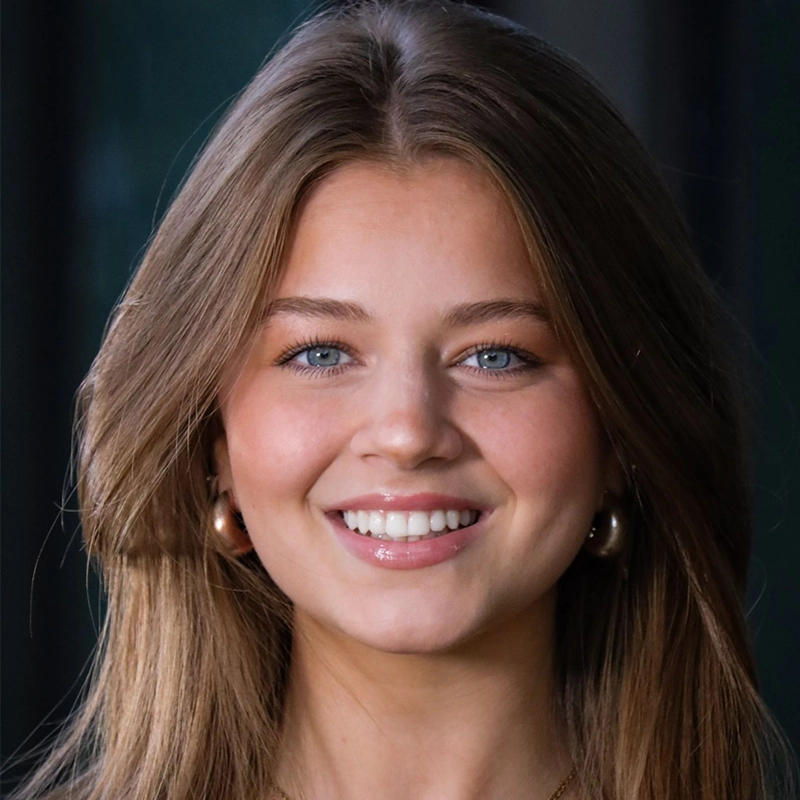A young woman with long, light brown hair and blue eyes smiles at the camera. She is wearing gold hoop earrings and natural makeup. The background is blurred and dark, making her face the focus of the image.