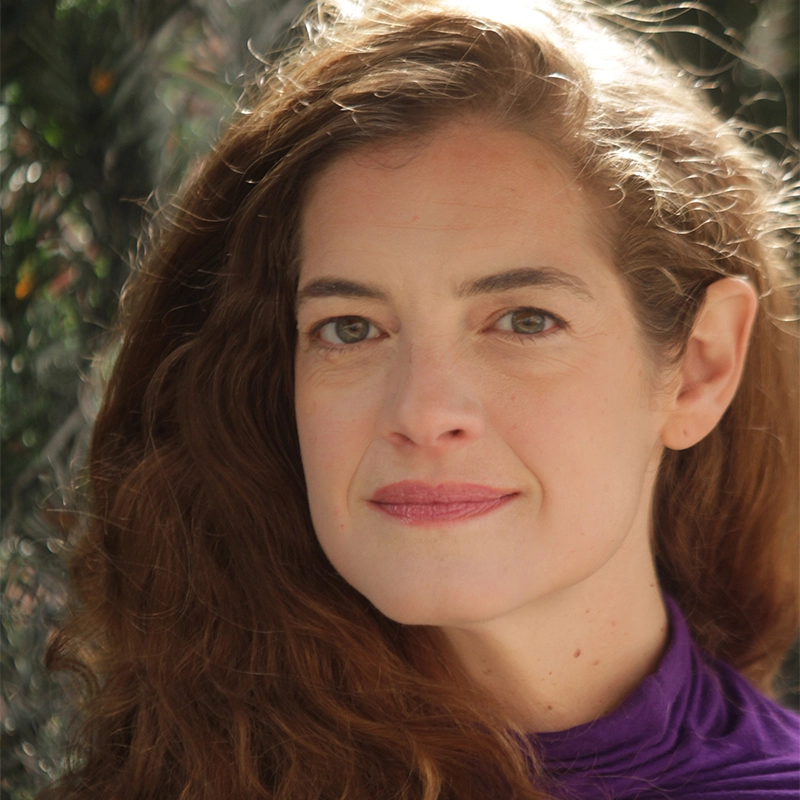 A portrait of Elyse Orecchio with long, wavy auburn hair looking directly at the camera. She has a gentle smile, hazel eyes, and is wearing a high-necked purple top, set against a softly blurred background of green foliage illuminated by dappled sunlight.