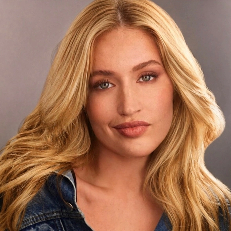 A headshot of a blonde woman with shoulder-length wavy hair and green eyes. She has a neutral expression and is wearing a denim jacket against a simple, light grey background.