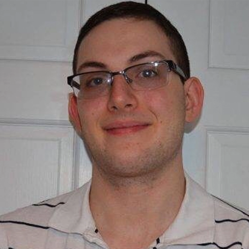 Tyler Cirulnick is a smiling man with short brown hair, glasses, and a white collared shirt with thin dark horizontal stripes, positioned in front of a white paneled door.