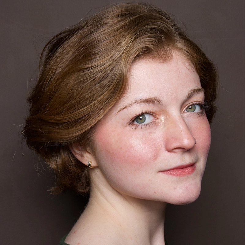 A young woman with fair skin, blue eyes, and short light brown hair looks slightly over her shoulder at the camera. She wears a small gold hoop earring and a dark top against a plain brown background. Her expression is calm and neutral.